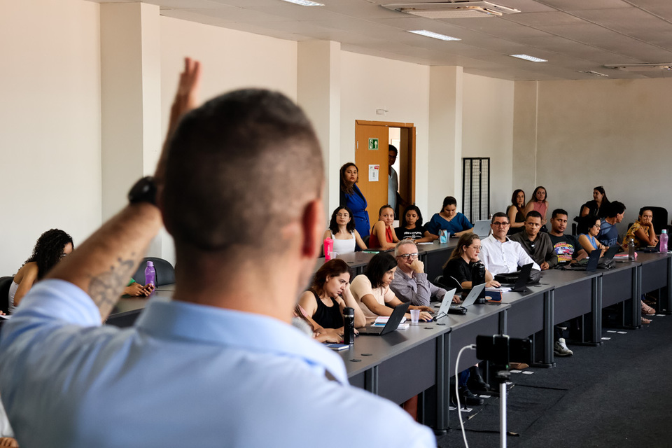 Homem branco ao microfone visto de costa com auditório cheio